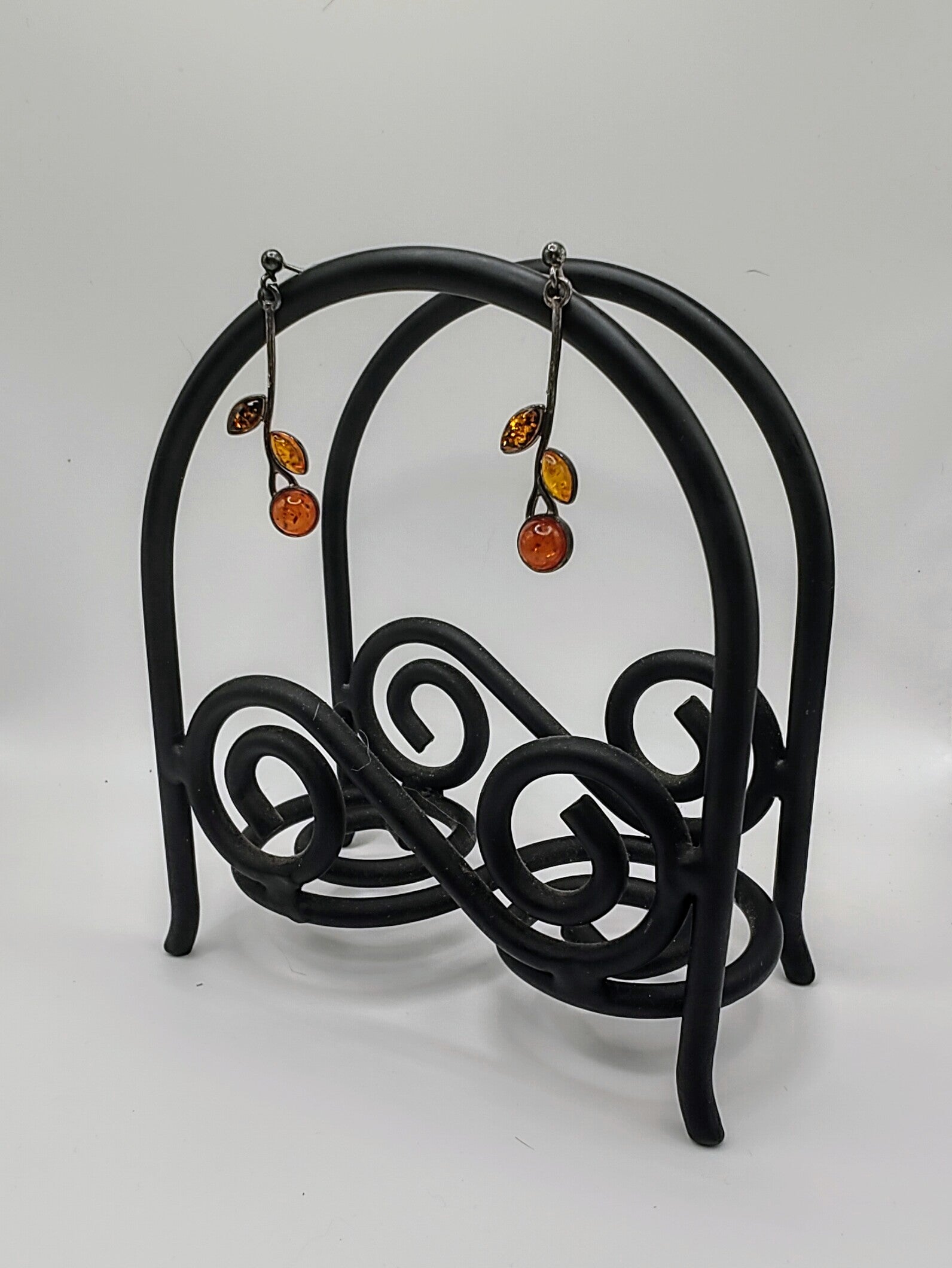 Amber Earrings hanging from a black scrollwork metal stand, showing their long silver vine shape with glowing amber stones.