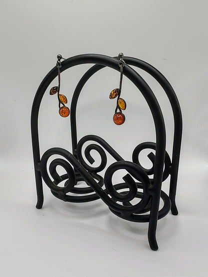 Amber Earrings hanging from a black scrollwork metal stand, showing their long silver vine shape with glowing amber stones.