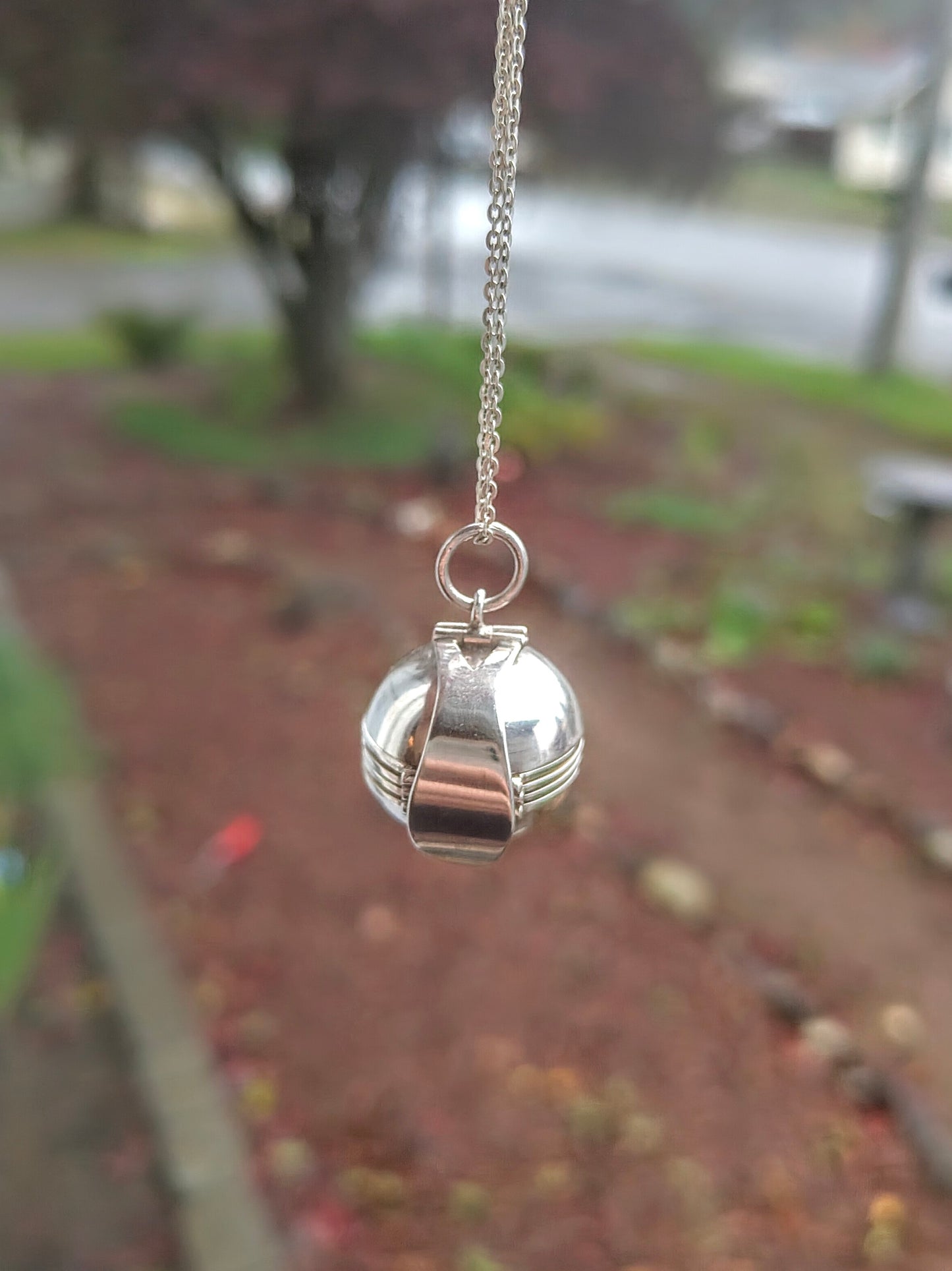 Sterling silver orb locket from the World Within Necklace hanging from a chain with a blurred garden background behind it.
