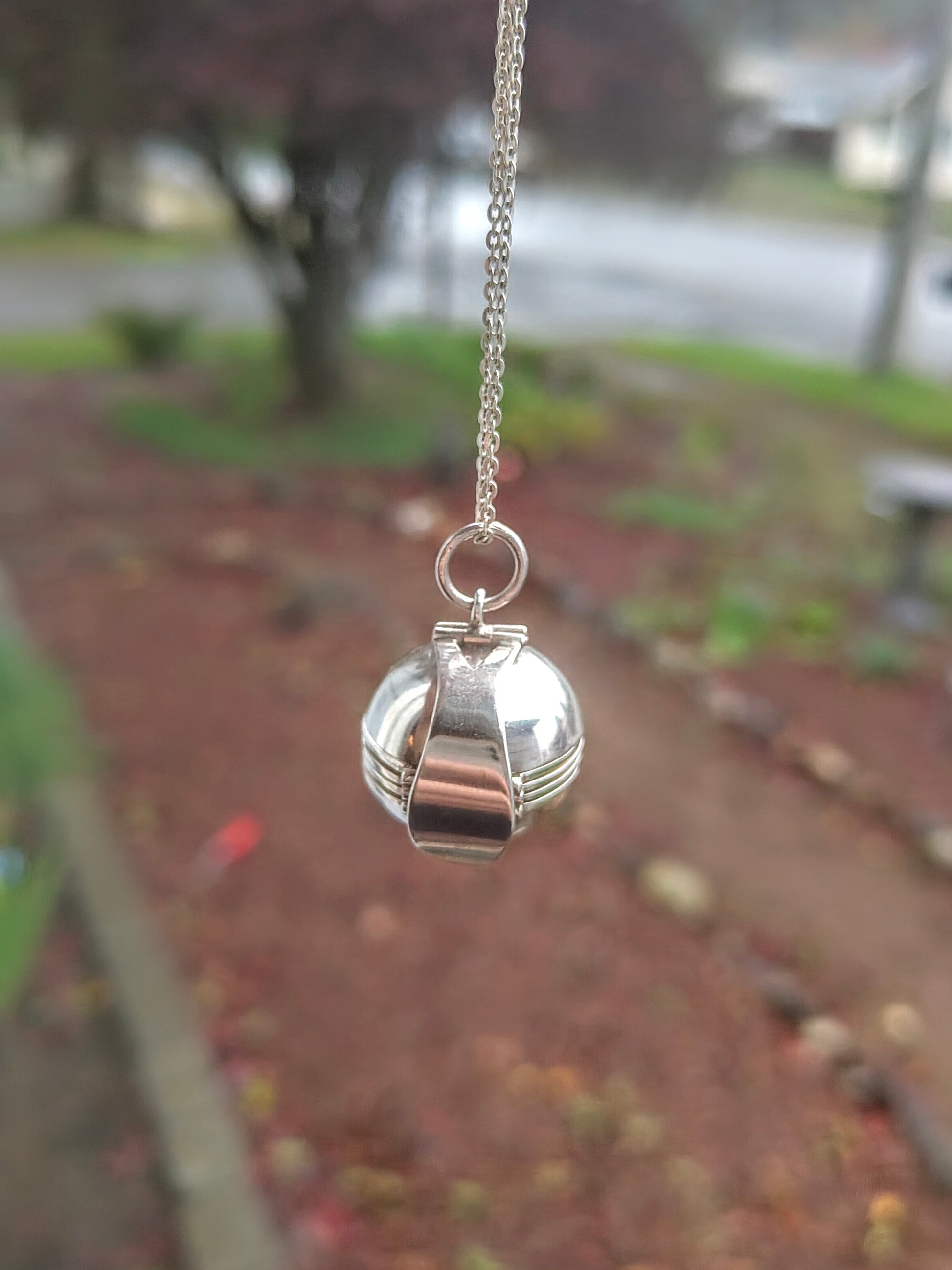 Sterling silver orb locket from the World Within Necklace hanging from a chain with a blurred garden background behind it.